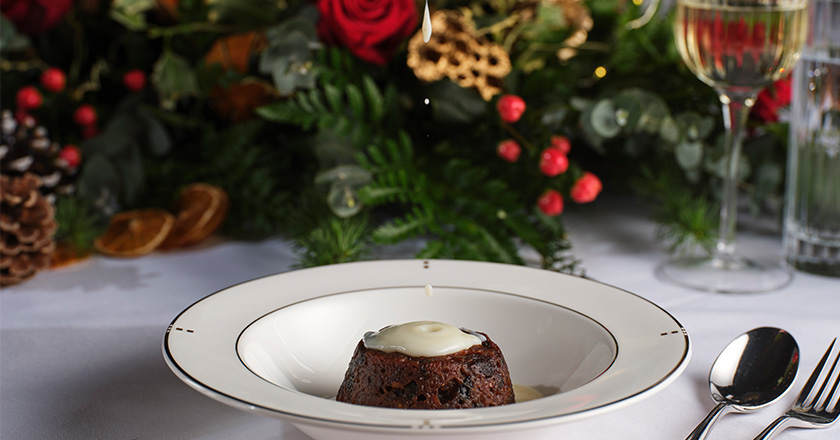 A Christmas pudding with white sauce on a white plate, and a Christmas foliage to decorate in background