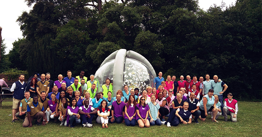 Large group of people wearing colourful bibs posing in front of an inflatable transparent cylinder, imitating the 'crystal maze' from TV.