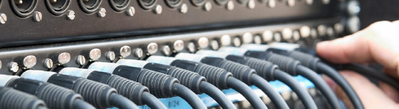 Electrical leads going into the back of a piece of audio visual equipment, with hand to the right plugging one more in.