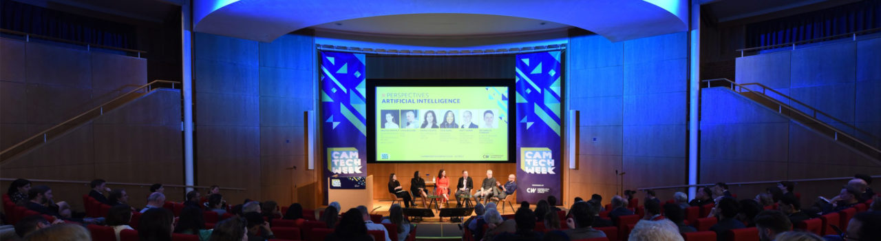 Photograph of the Francis Crick Auditorium stage decorated with blue lights and banners for Cam Tech Week, also showing panel discussion of six people.