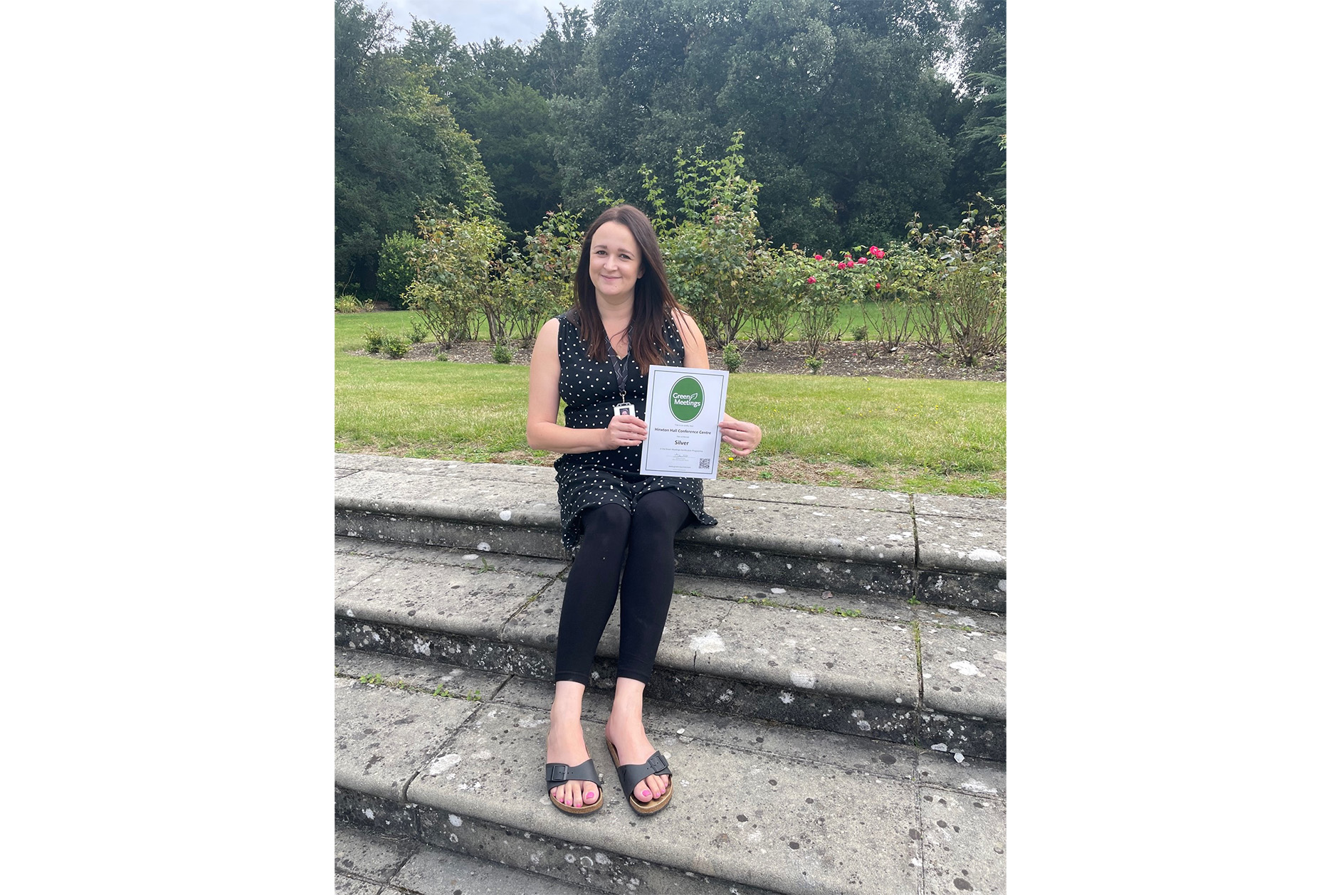 Kelly Butler, Business and Events Senior Manager, sat on steps on Hinxton Hall lawns holding Green Tourism award certificate