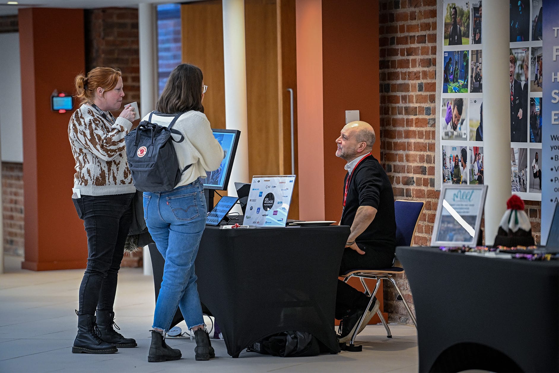 Ben McDade sat at desk speaking to two visitors to his exhibition stand, with examples of his photos behind him