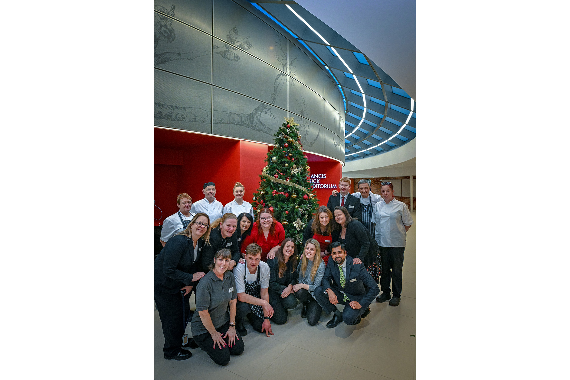 The teams behind the Hinxton Hall Open Afternoon - events and kitchen teams - standing in front of the Christmas tree and the Francis Crick Auditorium