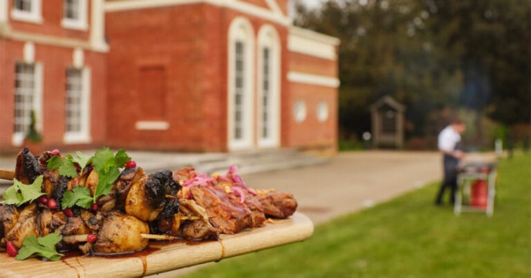 Wooden board with BBQ food on in foreground, blurred in background in chef at BBQ and part of Hinxton Hall.