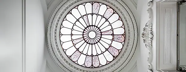 A round lantern window with a few remaining original glass panels in purple-y grey featuring fleur de Lys like images. The rest are plain opaque white glass. Surrounded by white painted ceiling with some Rococo details.