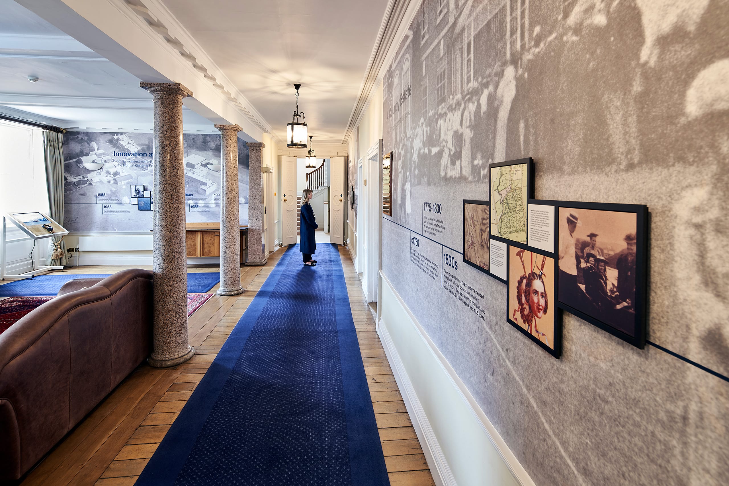Hinxton Hall Foyer, with wall on right covered in large black and white photograph and with smaller frames with historical information on top. To the left, two granite columns support ceiling. In between the two, a long thin blue carpet, with an exhibition visitor dressed in black, at the end. Hinxton Hall Foyer, with wall on right covered in large black and white photograph and with smaller frames with historical information on top. To the left, two granite columns support ceiling. In between the two, a long thin blue carpet, with an exhibition visitor dressed in black, at the end.