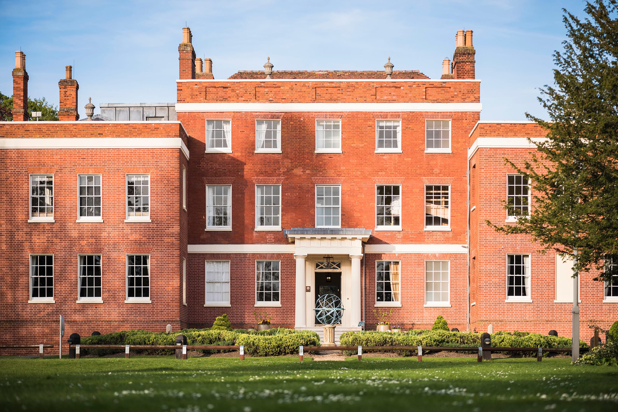 Mid shot of the front of Hinxton Hall, red brick with white mantels and large white sash windows. Black front door with white porch. Mid shot of the front of Hinxton Hall, red brick with white mantels and large white sash windows. Black front door with white porch.