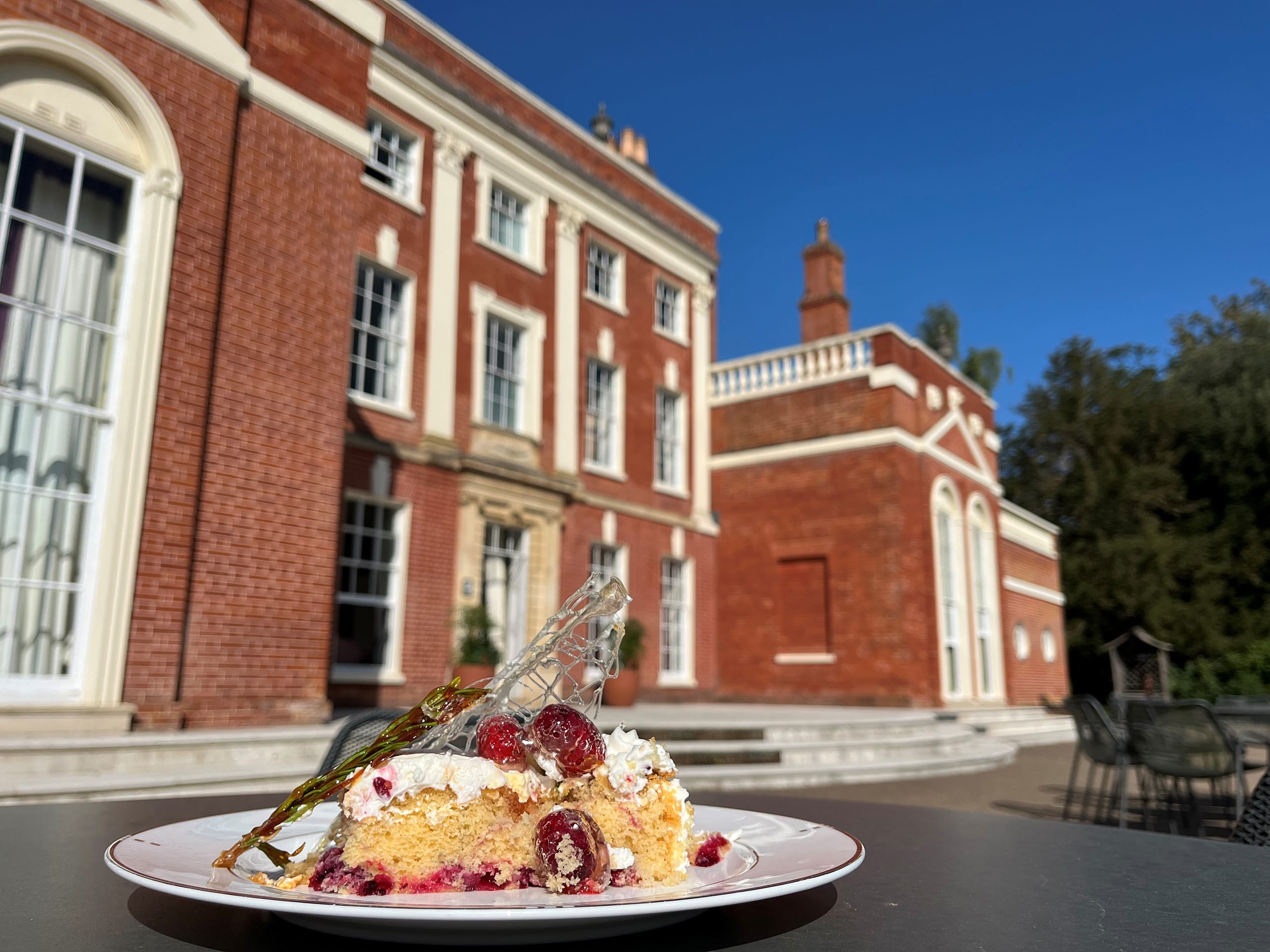 A slice of mulberry and lemon cake with icing on top, set on a table outside Hinxton hall, visible in the background