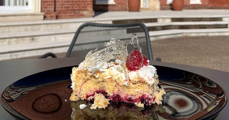 A slice of mulberry and lemon cake with icing on top, set on a table outside Hinxton hall, visible in the background.