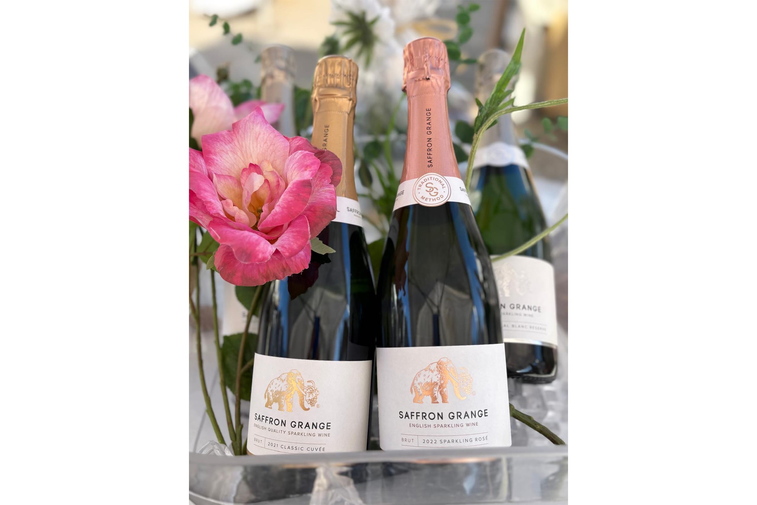 A bottle of Saffron Grange's 2021 Classic Cuv&eacute;e and of their 2022 Sparkling Ros&eacute; sitting in a wine bucket alongside a beautiful large pink flower.