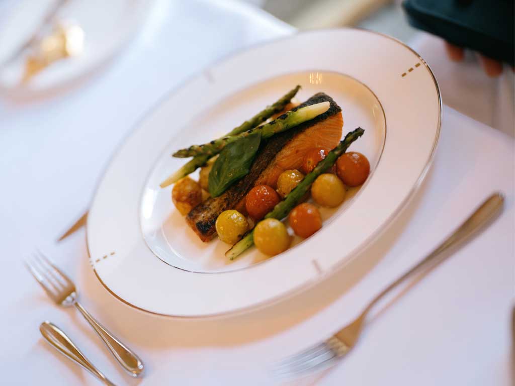 A plate of salmon with roasted vegetables on a white plate, with a white tablecloth, knife and fork.